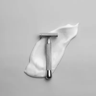 A luxurious men's grooming setup in a modern, moody bathroom. The product is resting on a wet, dark matte slate stone surface with natural textures. Scattered, realistic water droplets surround the product. In the blurred background, a hint of dark green ceramic wall tiles and a subtle wisp of steam. Morning natural light creates soft, high-contrast reflections and perfect highlights on the metallic surface of the product. The atmosphere is clean, refreshing, and highly premium.