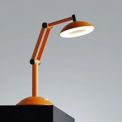 A sophisticated, creative late-night workspace. The product is placed on a premium walnut wood desk with a warm, natural grain. The lamp is turned on, casting a warm, inviting pool of light directly onto a partially unrolled architectural blueprint and a sleek metal fountain pen resting on the desk. The surrounding room is cast in moody, cinematic shadows, with the blurred silhouette of a bookshelf in the dark background. The composition conveys a quiet, focused, and cozy mid-century modern aesthetic.